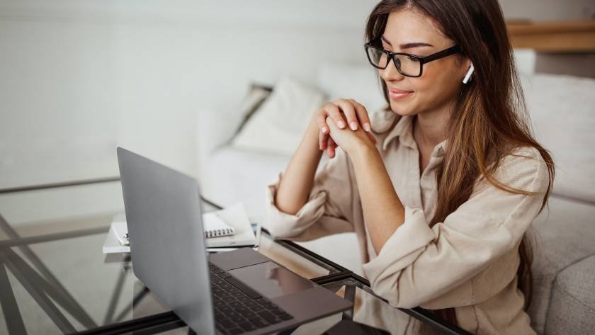 Smiling millennial mixed race woman in wireless headphones and glasses has video call on laptop